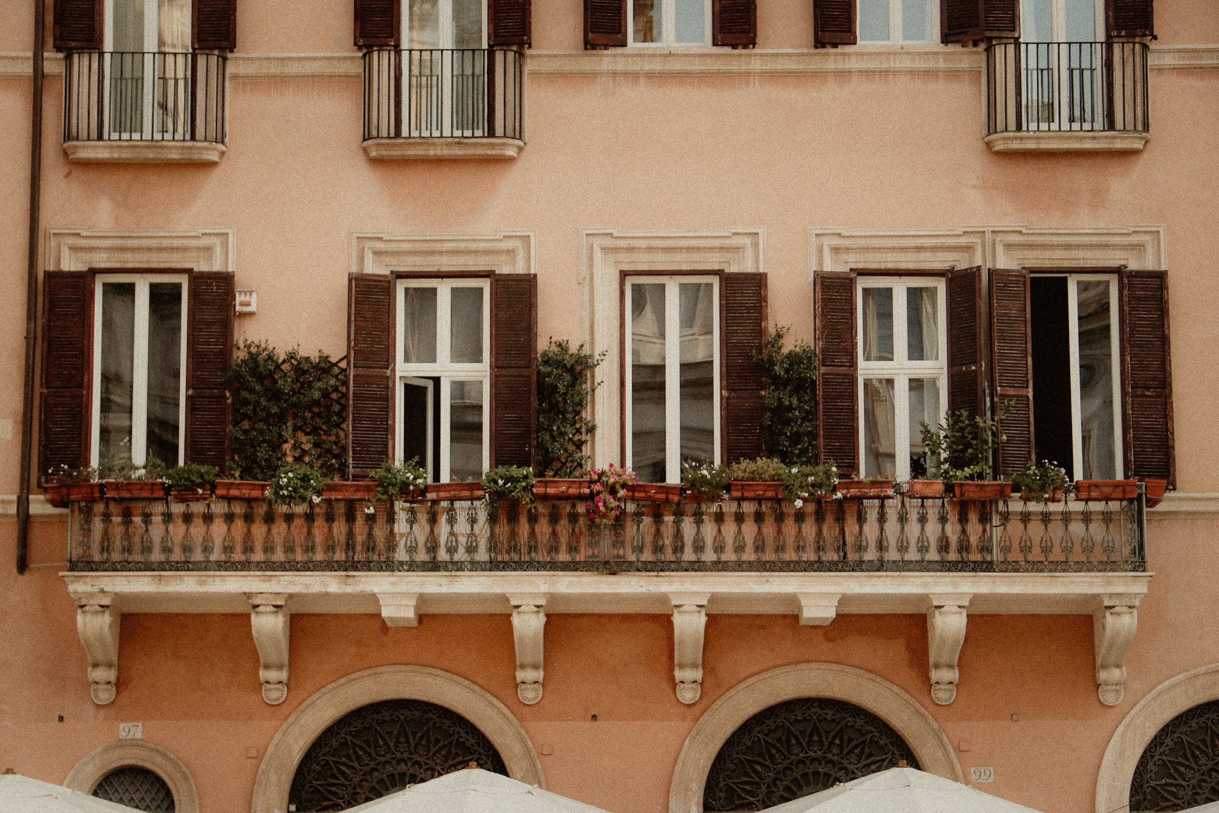 A long Roman balcony with dark shutters, pale stone and flower boxes