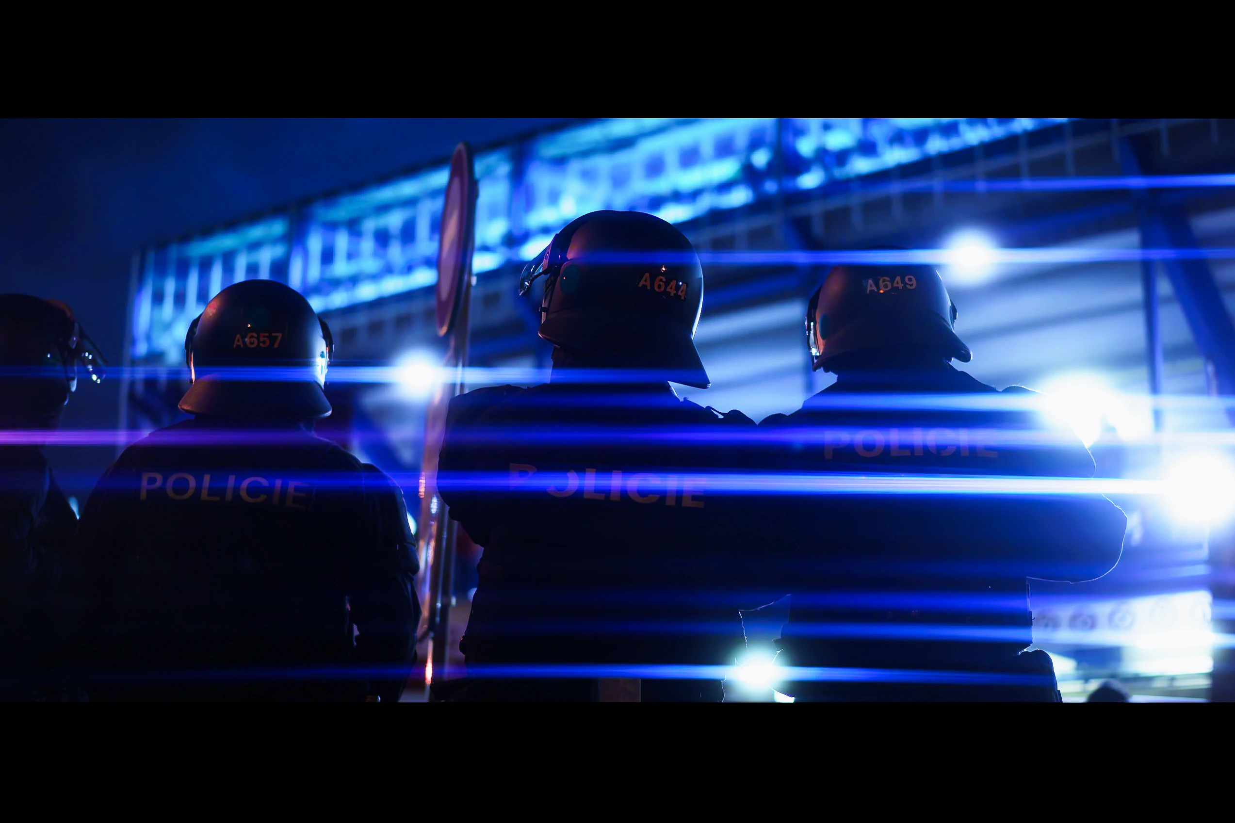 Four Czech police officers seen from behind as blue emergency lights form horizontal streaks across the frame