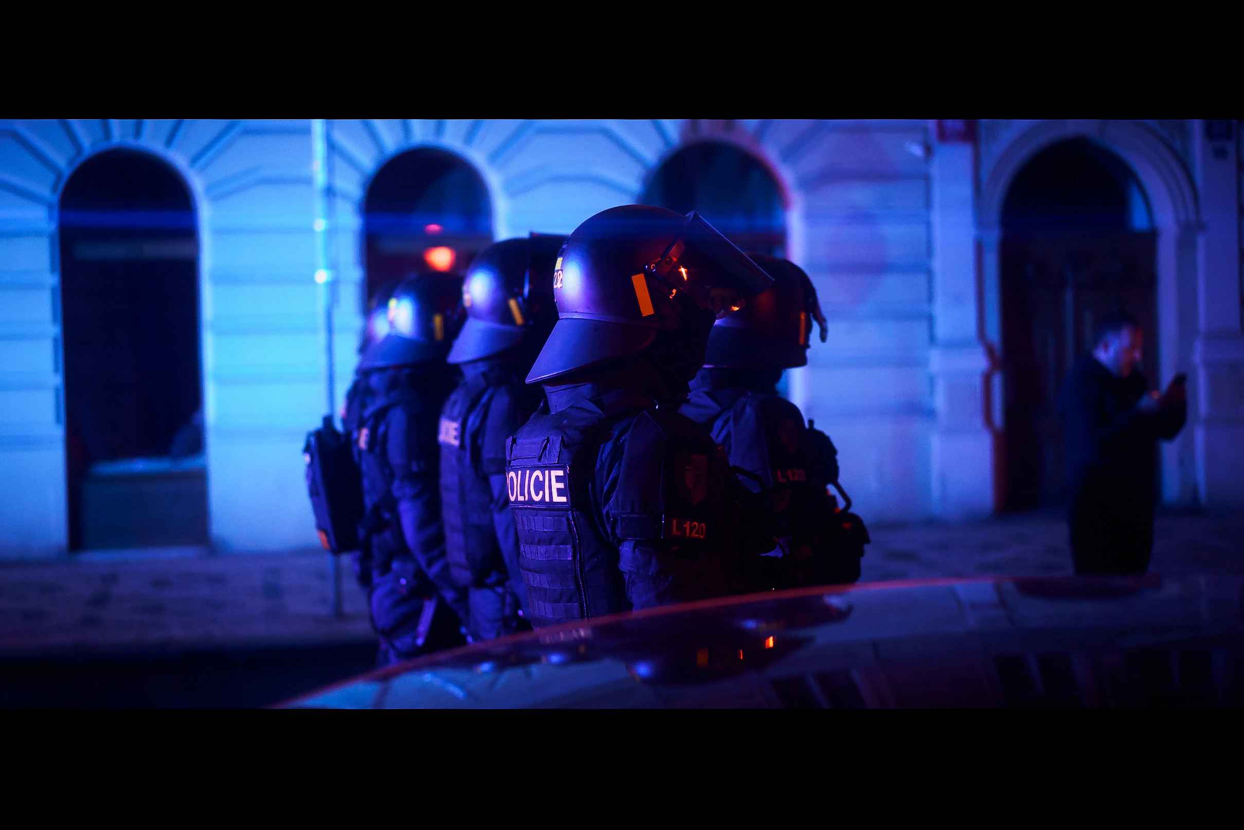 A line of Czech police officers in riot gear under blue light on a Prague street