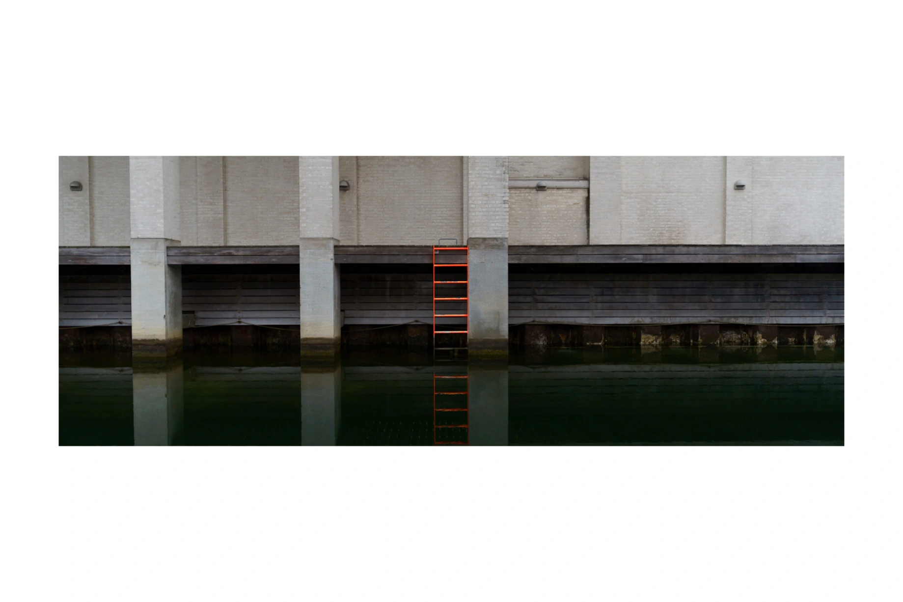 A red ladder reflected in dark water below an industrial facade
